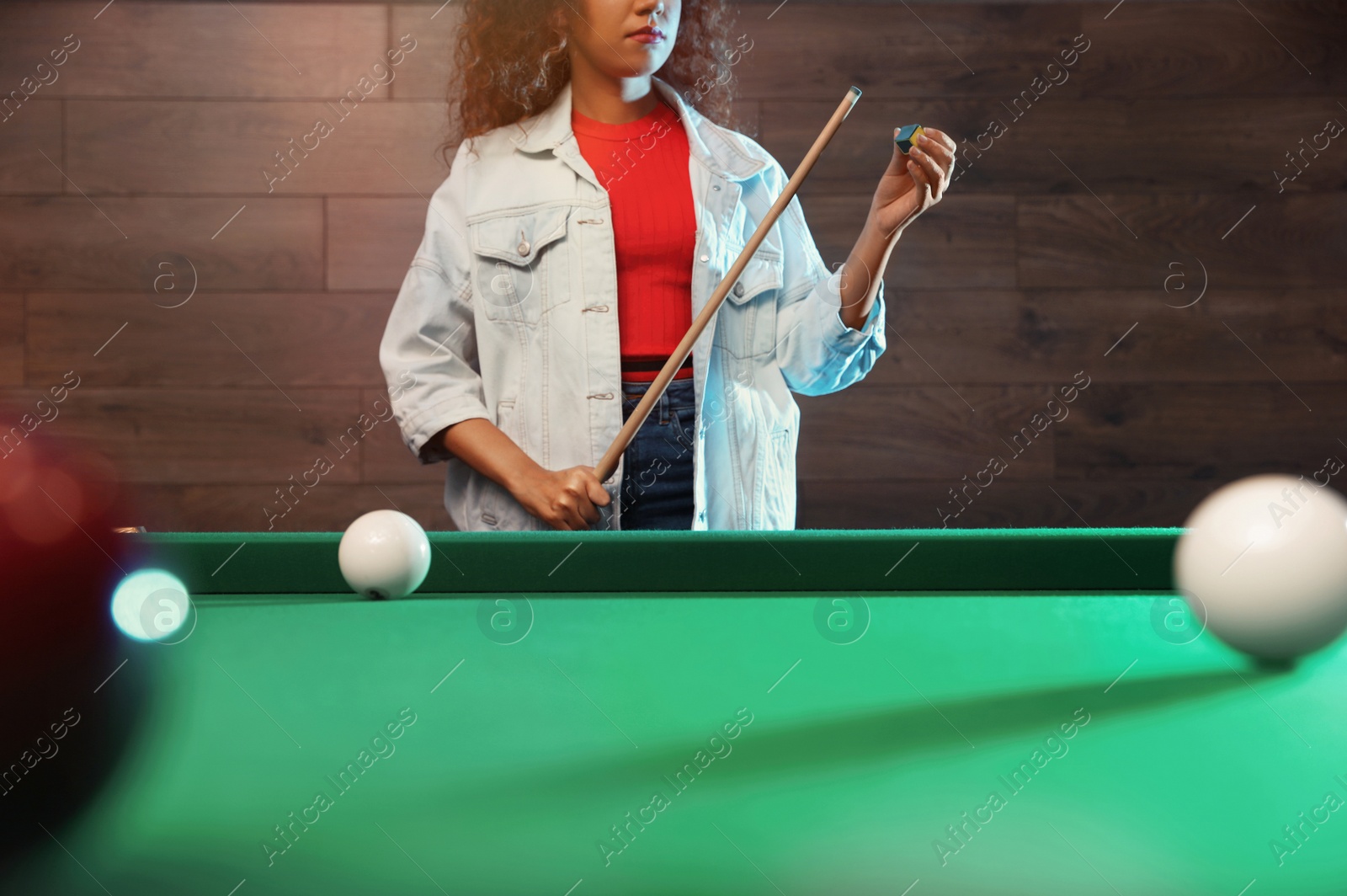 Photo of Young African-American woman chalking cue near billiard table indoors, closeup