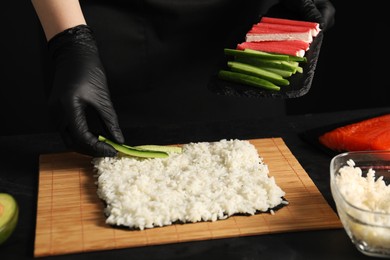 Chef in gloves putting cucumber onto unwrapped sushi roll at dark table, closeup