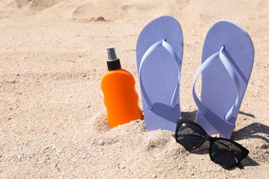 Photo of Stylish flip flops, sunglasses and sun protection spray on sand, space for text