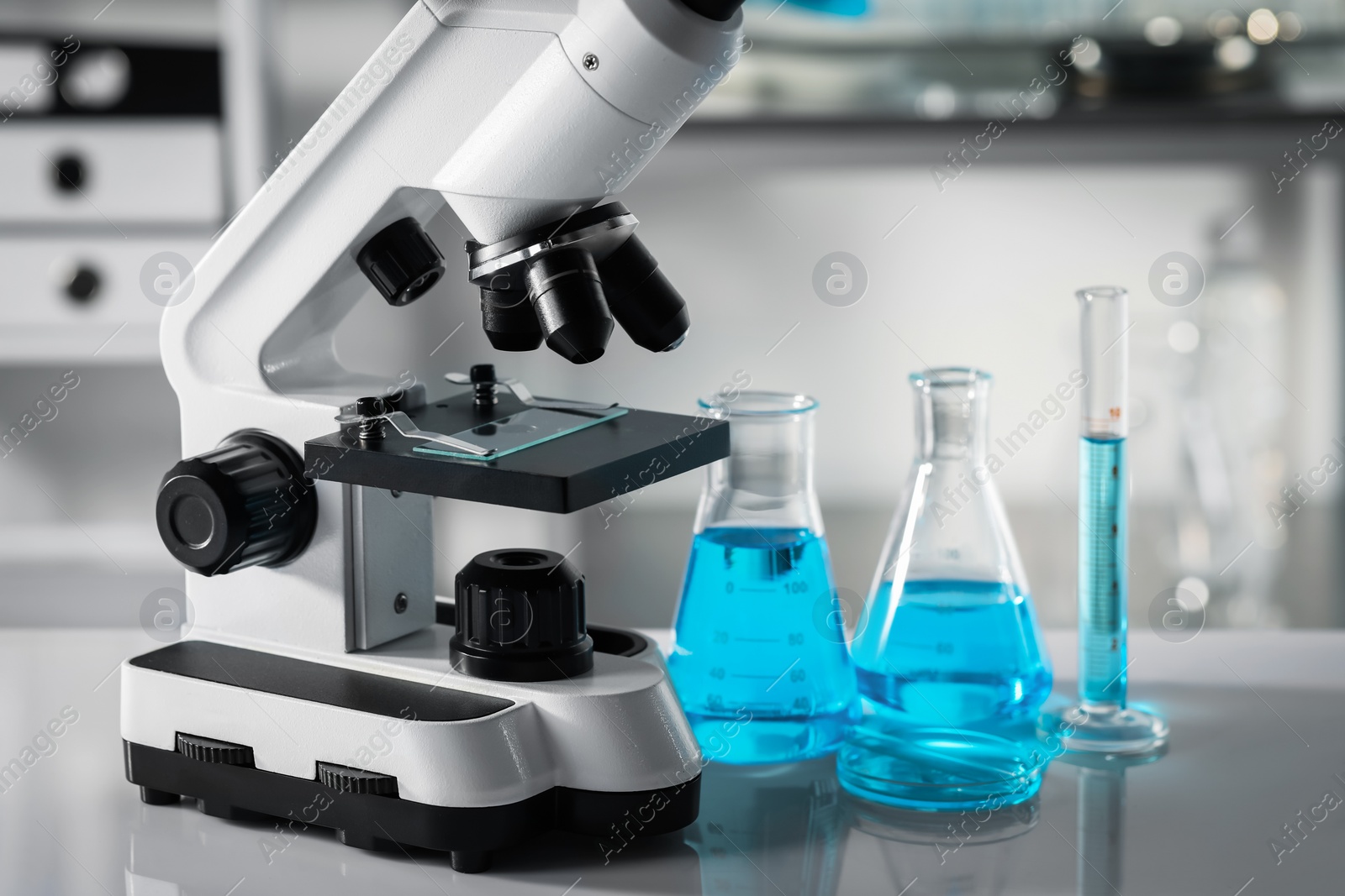 Photo of Different laboratory glassware with light blue liquid and microscope on table