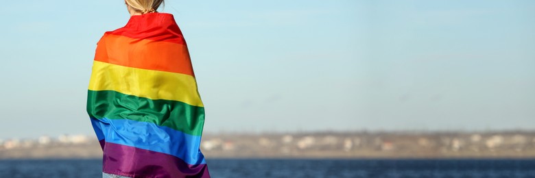 Woman wrapped in bright LGBT flag near river, back view with space for text. Banner design