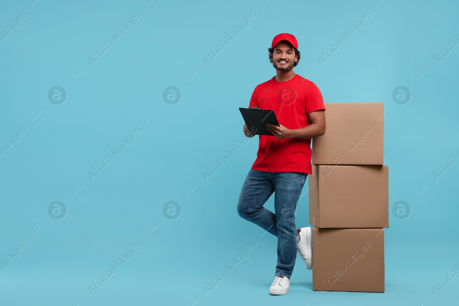 Photo of Happy young courier with clipboard and stack of parcels on light blue background, space for text