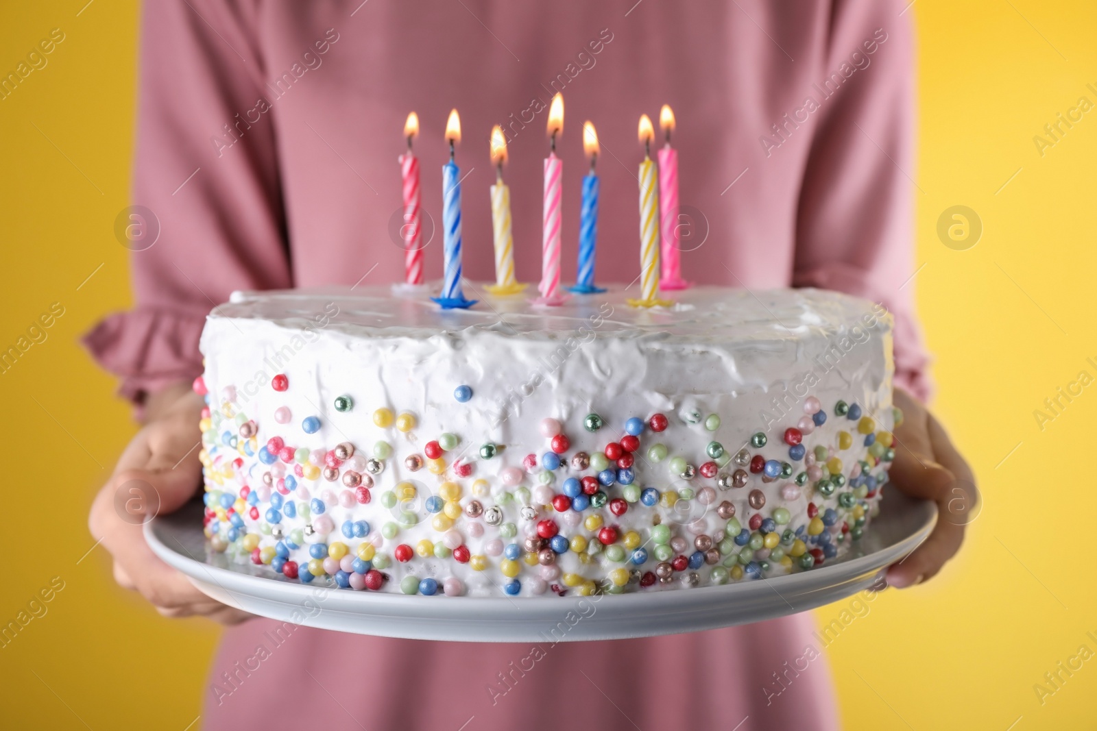 Photo of Woman holding birthday cake with burning candles on yellow background, closeup