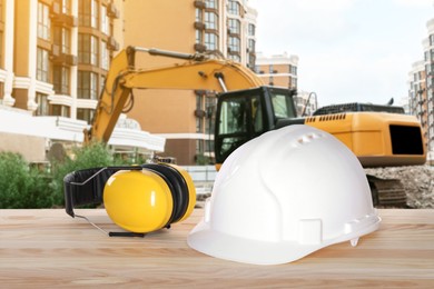 Image of Safety equipment. Hard hat and protective headphones on wooden surface near buildings and excavator outdoors