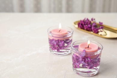 Glass holders with water, lilac flowers and burning candles on light table, space for text