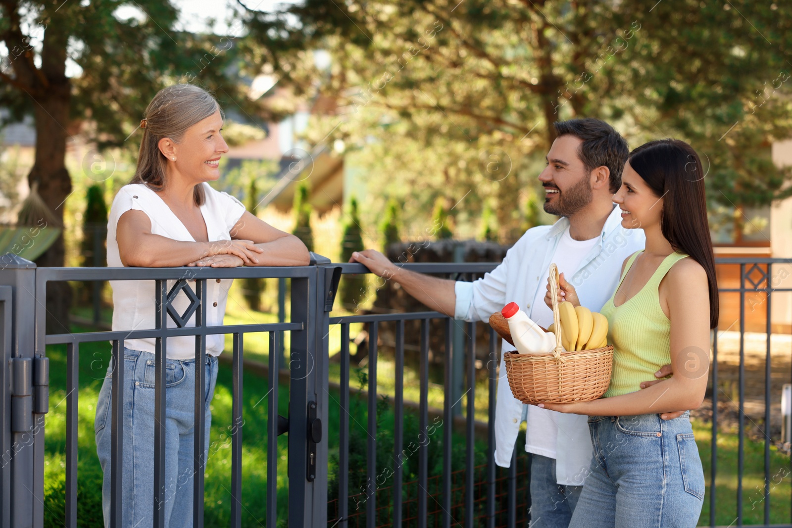 Photo of Friendly relationship with neighbours. Young couple with wicker basket of products treating senior woman near fence outdoors