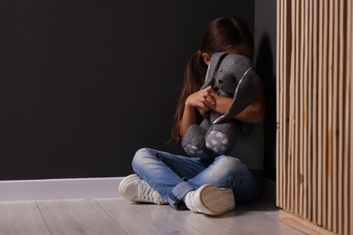 Child abuse. Upset little girl with toy bunny sitting on floor near gray wall indoors, space for text