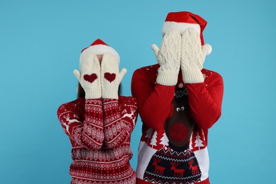 Photo of Young couple in Christmas sweaters and Santa hats covering faces with hands in knitted mittens on light blue background