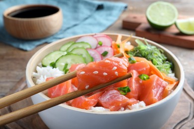 Photo of Wooden chopsticks with piece of salmon over delicious poke bowl on table, closeup