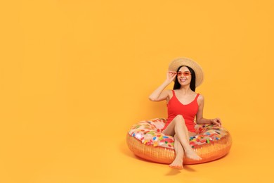 Photo of Happy young woman with beautiful suntan, hat and sunglasses on inflatable ring against orange background, space for text