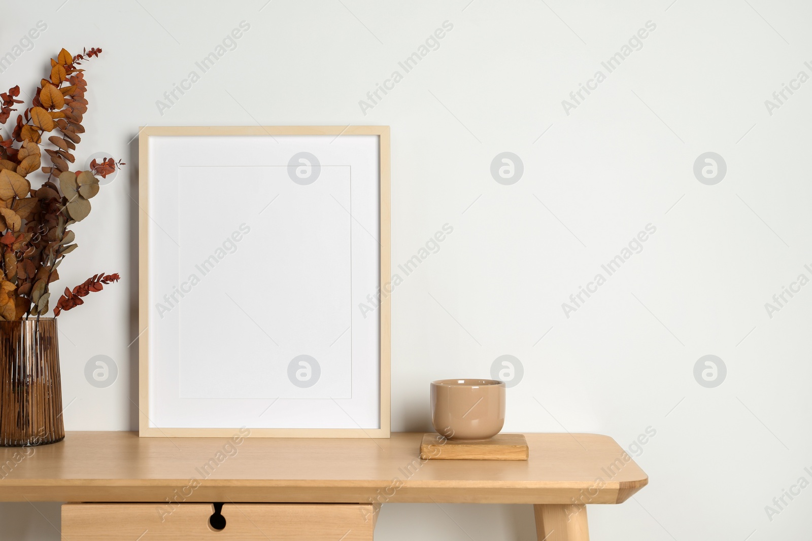 Photo of Empty photo frame, cup and vase with dry decorative leaves on wooden table. Mockup for design