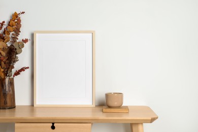 Photo of Empty photo frame, cup and vase with dry decorative leaves on wooden table. Mockup for design