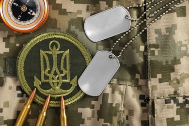 Photo of MYKOLAIV, UKRAINE - SEPTEMBER 26, 2020: Metal military ID tags near Ukrainian army patch, bullets and compass on camouflage background, flat lay
