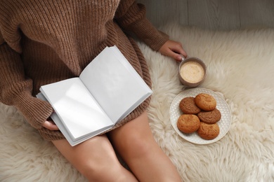 Photo of Young woman with cup of coffee reading book on rug at home, above view. Winter atmosphere