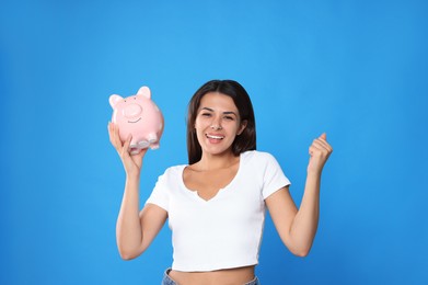 Happy young woman with piggy bank on light blue background