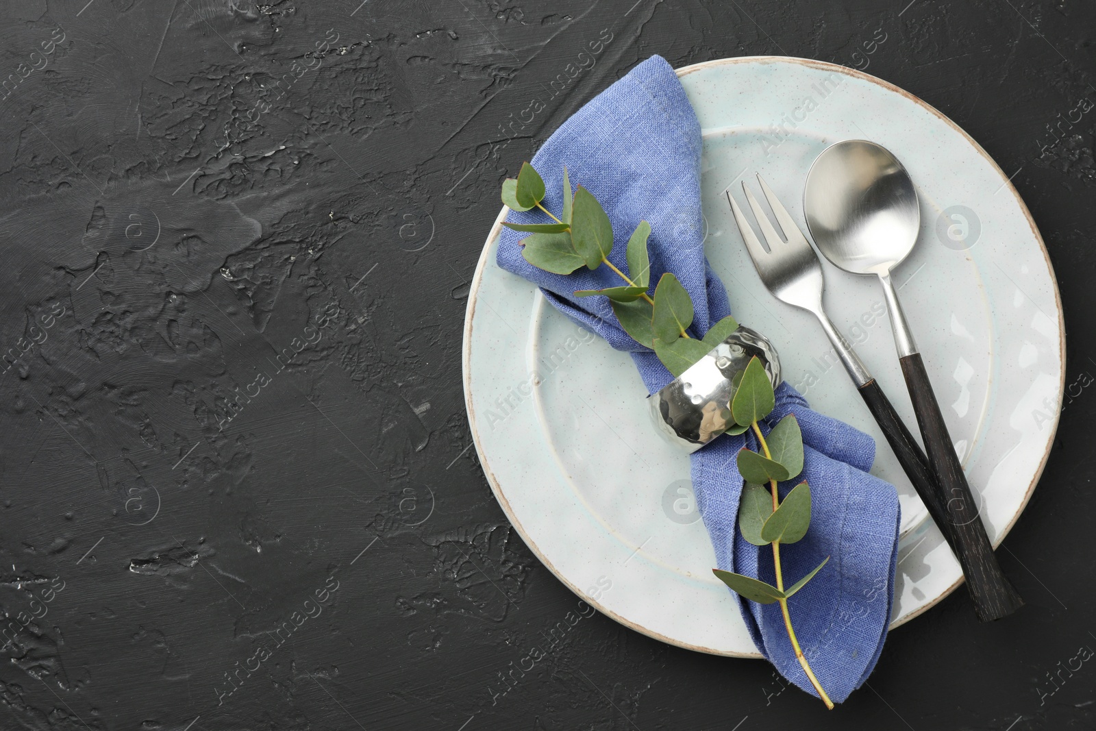 Photo of Stylish setting with cutlery, eucalyptus branch, napkin and plate on dark textured table, top view. Space for text