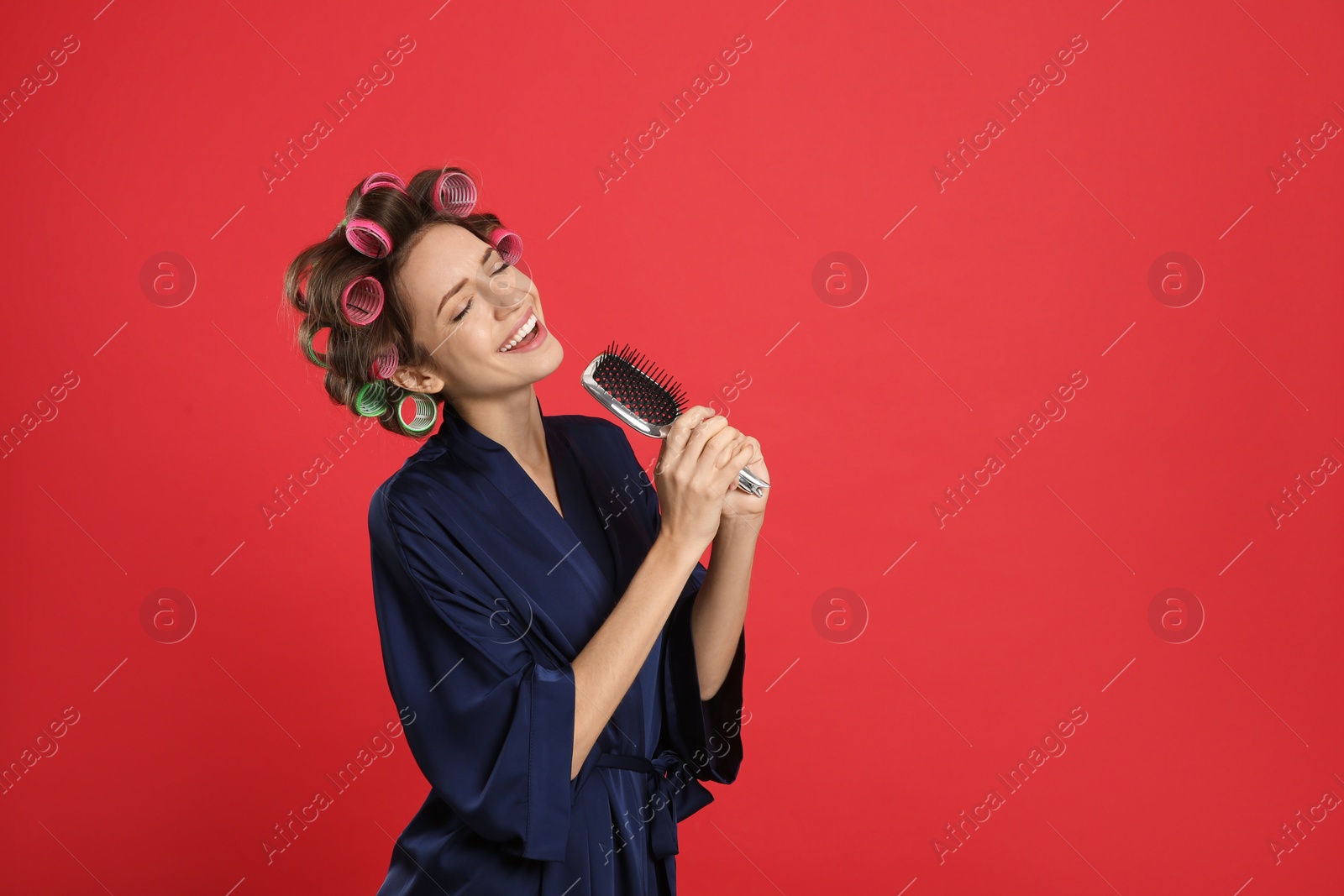 Photo of Beautiful young woman in silk bathrobe with hair curlers singing into hairbrush on red background. Space for text