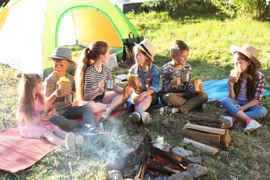 Photo of Little children eating sandwiches near bonfire and tent. Summer camp