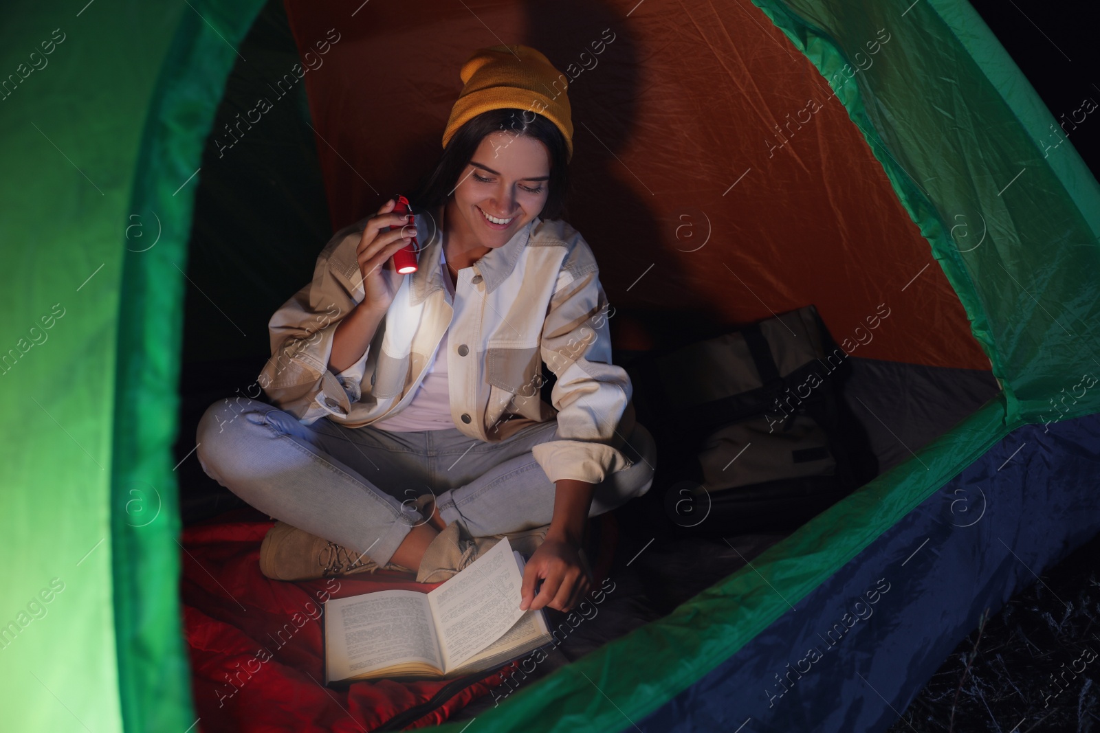 Photo of Young woman with flashlight reading book in tent at night