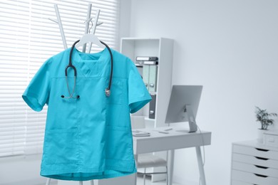 Turquoise medical uniform and stethoscope hanging on rack in clinic. Space for text