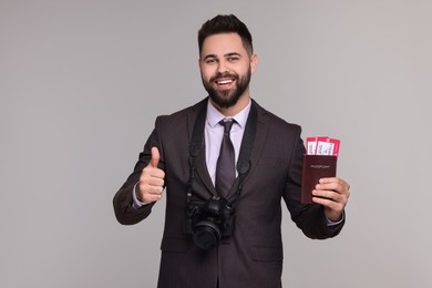 Happy businessman with passport, tickets and camera showing thumb up on grey background