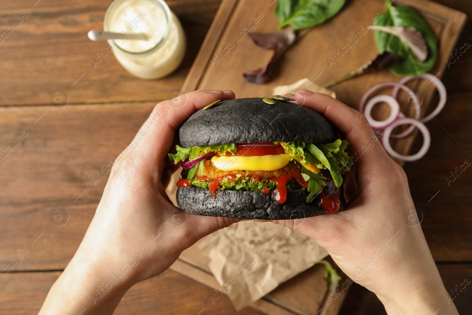 Photo of Woman holding tasty black vegetarian burger over table, top view