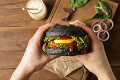 Woman holding tasty black vegetarian burger over table, top view