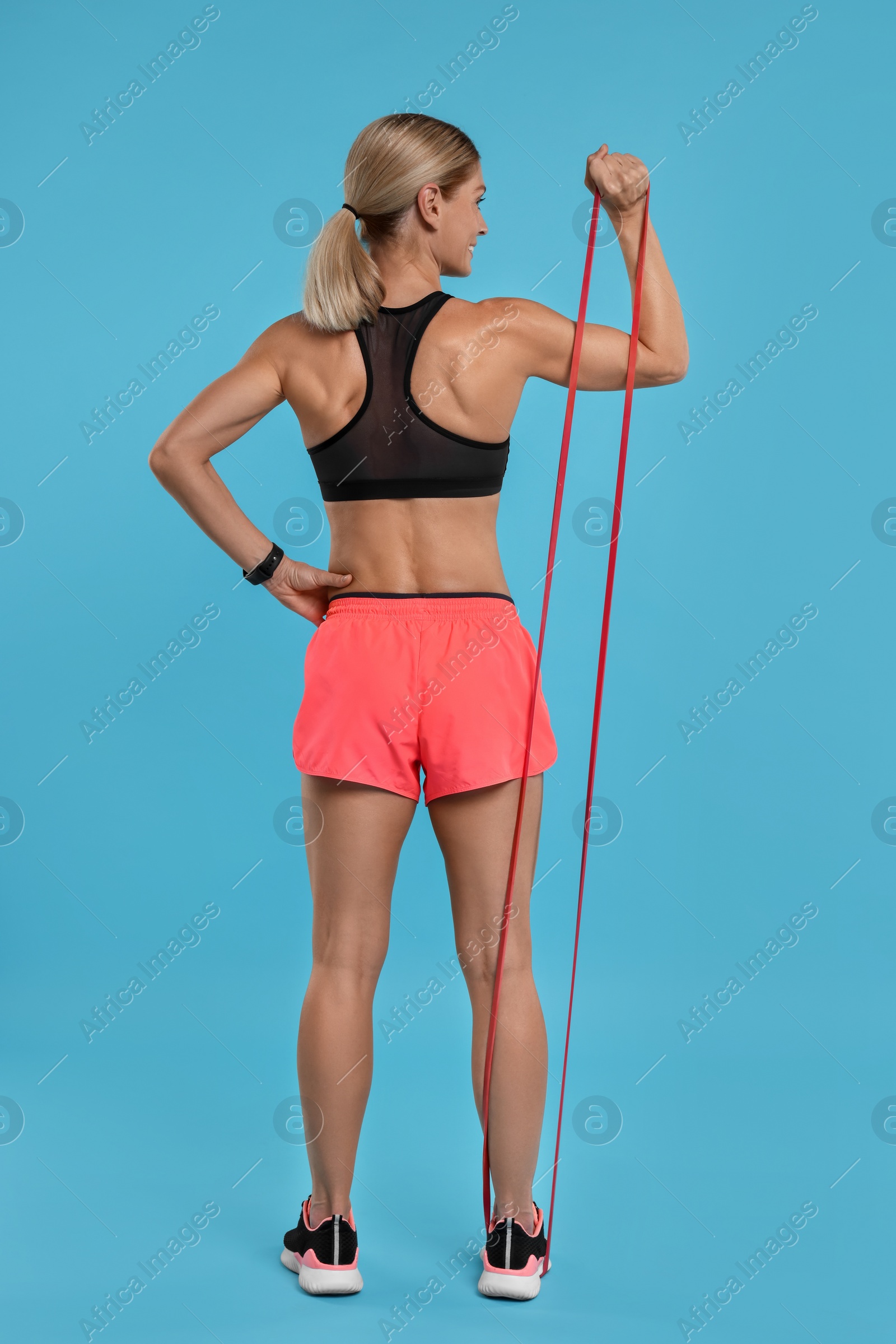 Photo of Woman exercising with elastic resistance band on light blue background, back view