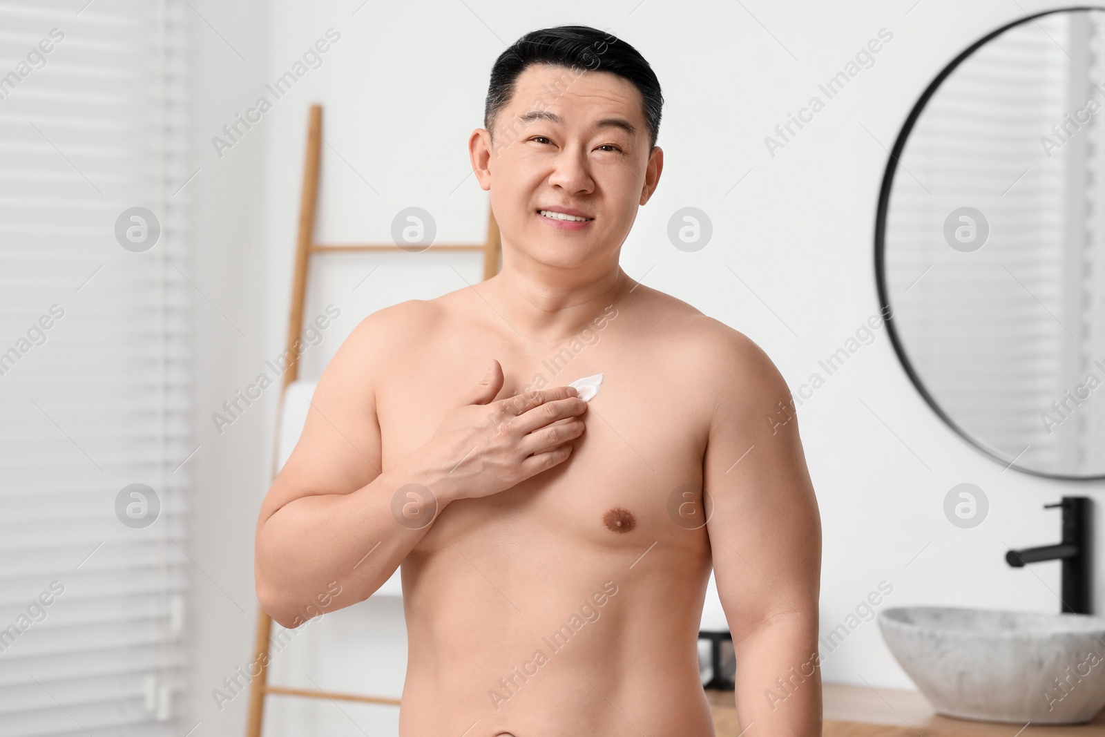 Photo of Handsome man applying body cream onto his chest in bathroom