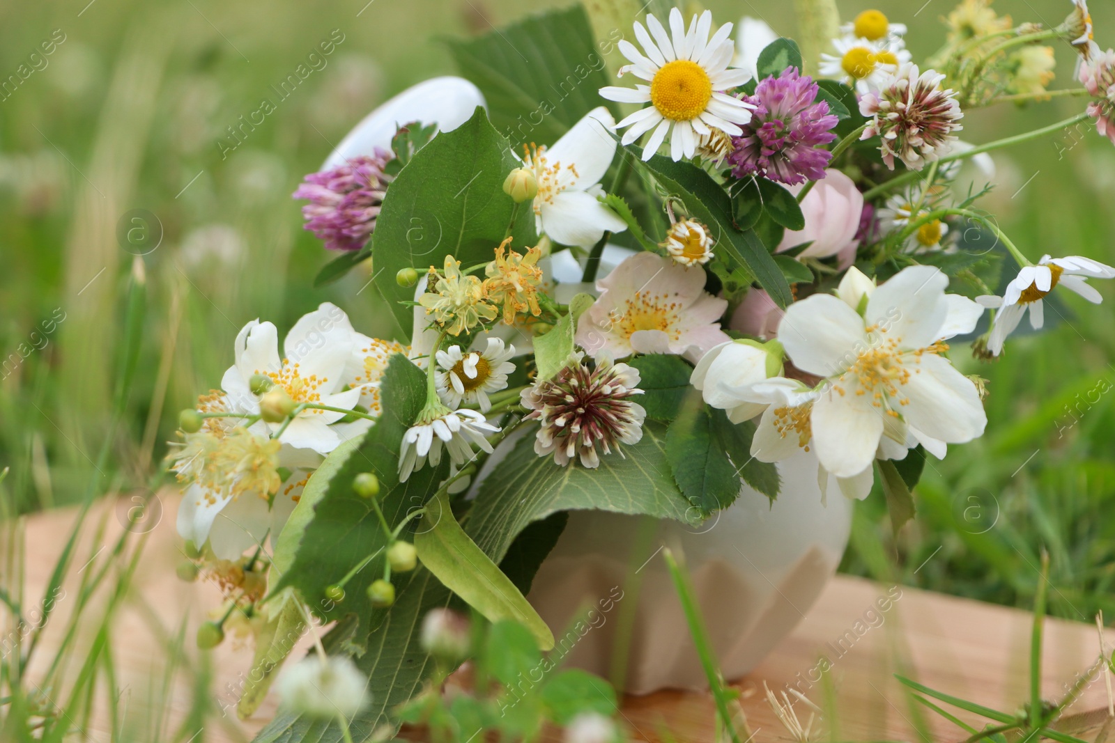 Photo of Ceramic mortar with pestle, different wildflowers and herbs on wooden board in meadow, closeup