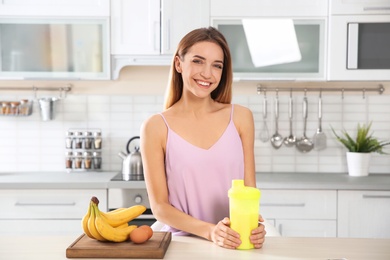 Young woman holding bottle of protein shake at table with ingredients in kitchen