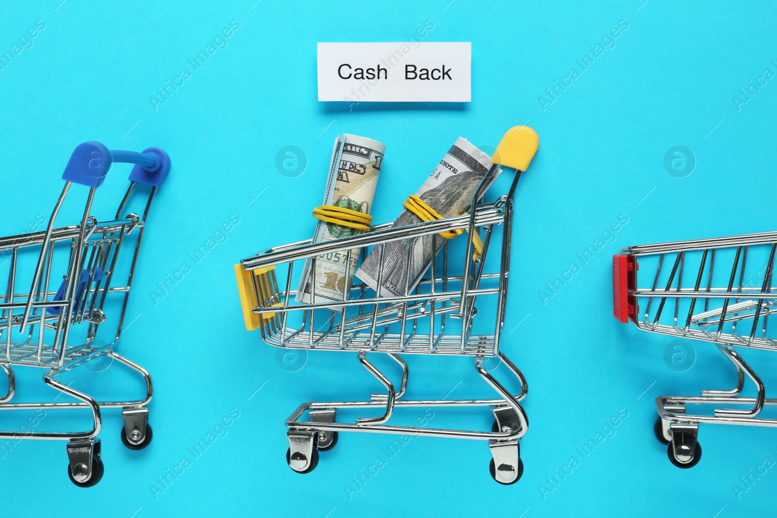 Photo of Card with word Cashback, rolled dollar banknotes and shopping carts on light blue background, flat lay