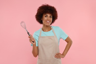 Photo of Happy young woman in apron holding whisk on pink background