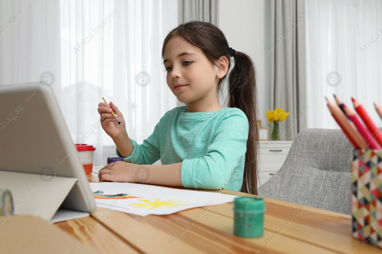 Photo of Little girl drawing on paper with paints at online lesson indoors. Distance learning
