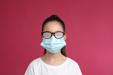Photo of Little girl with foggy glasses caused by wearing medical face mask on pink background. Protective measure during coronavirus pandemic