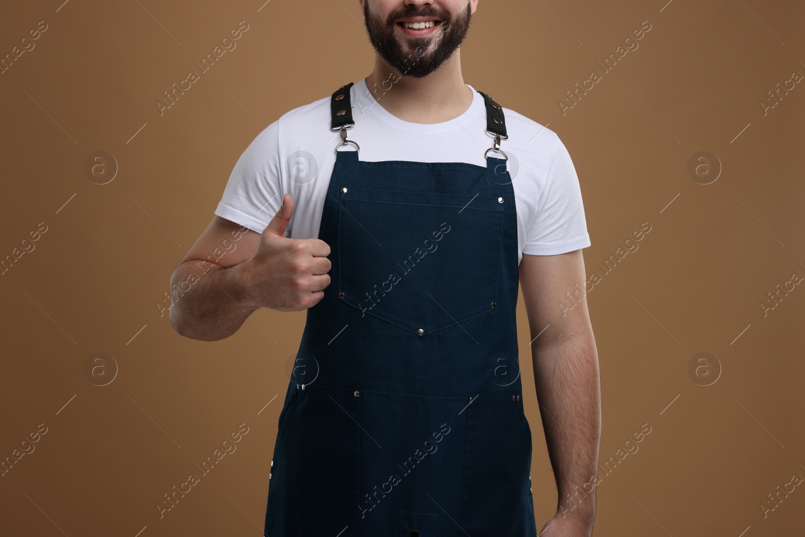 Photo of Smiling man in kitchen apron showing thumb up on brown background, closeup. Mockup for design
