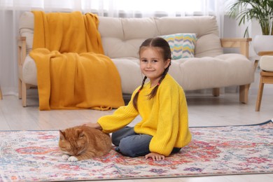 Photo of Smiling little girl petting cute ginger cat on carpet at home