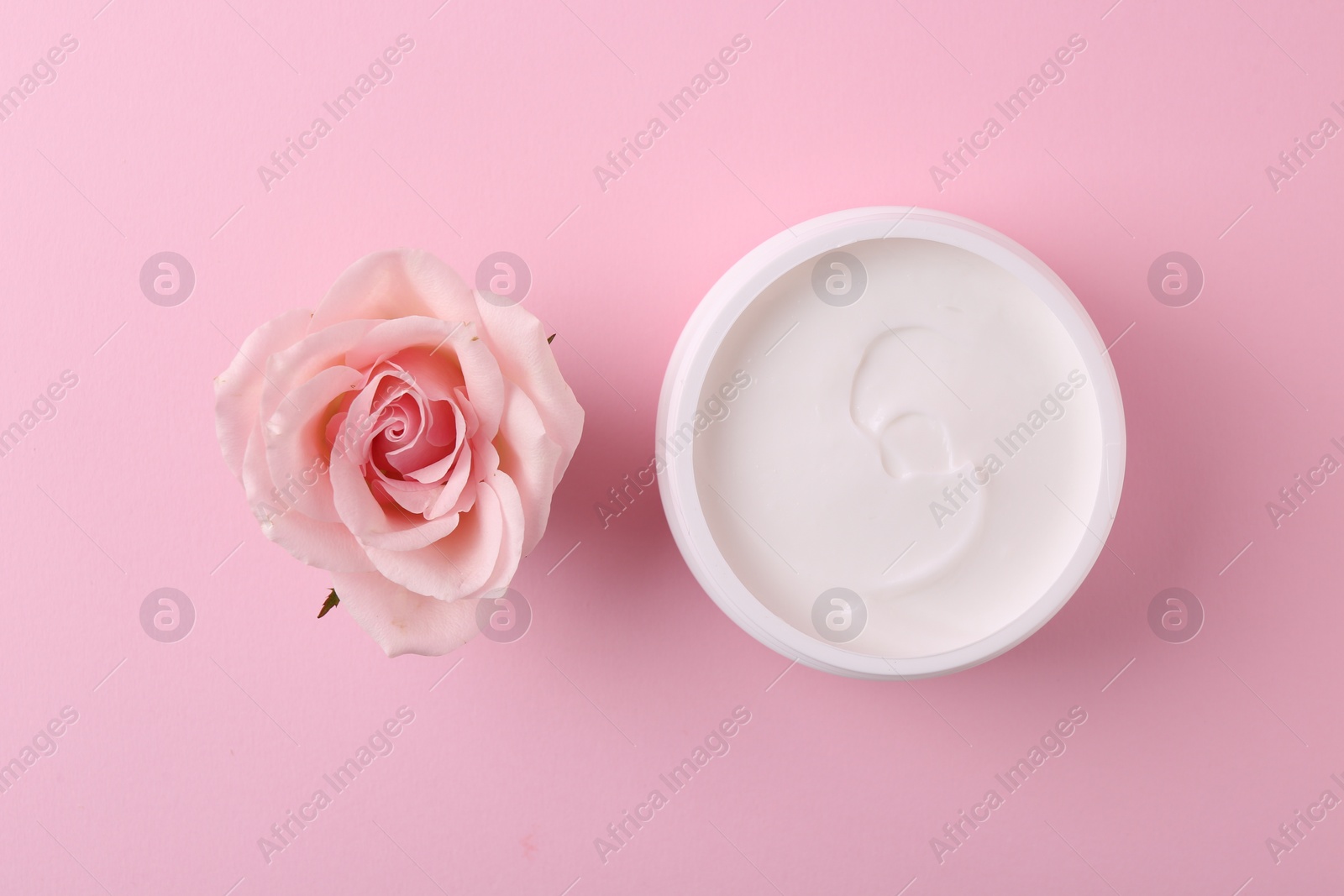 Photo of Moisturizing cream in open jar and rose flower on pink background, flat lay. Body care product