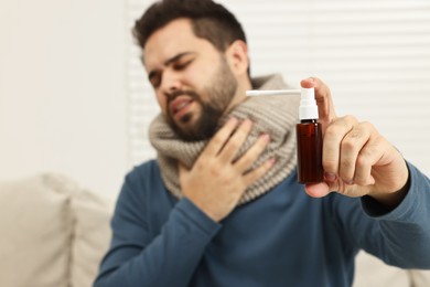 Young man with scarf holding throat spray indoors, selective focus