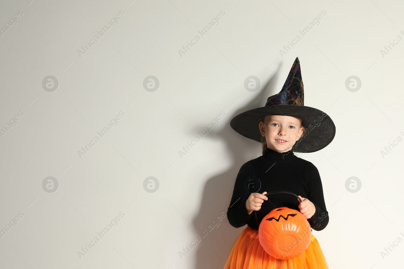 Photo of Cute little girl with pumpkin candy bucket wearing Halloween costume on light background. Space for text