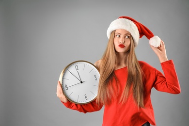 Young beautiful woman in Santa hat holding big clock on grey background. Christmas celebration