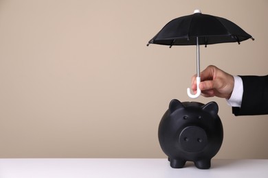 Woman holding small umbrella over piggy bank against beige background, closeup. Space for text