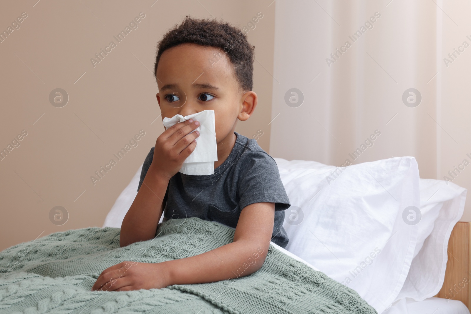 Photo of African-American boy with tissue blowing nose in bed indoors, space for text. Cold symptoms