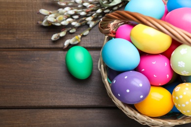 Photo of Colorful Easter eggs in wicker basket and willow branches on wooden table, flat lay. Space for text