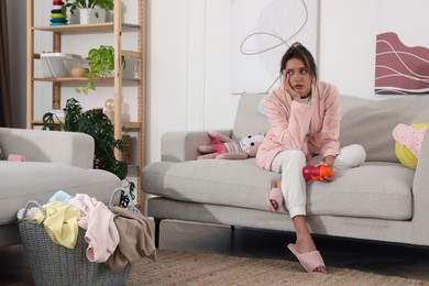 Photo of Tired young mother sitting on sofa in messy living room