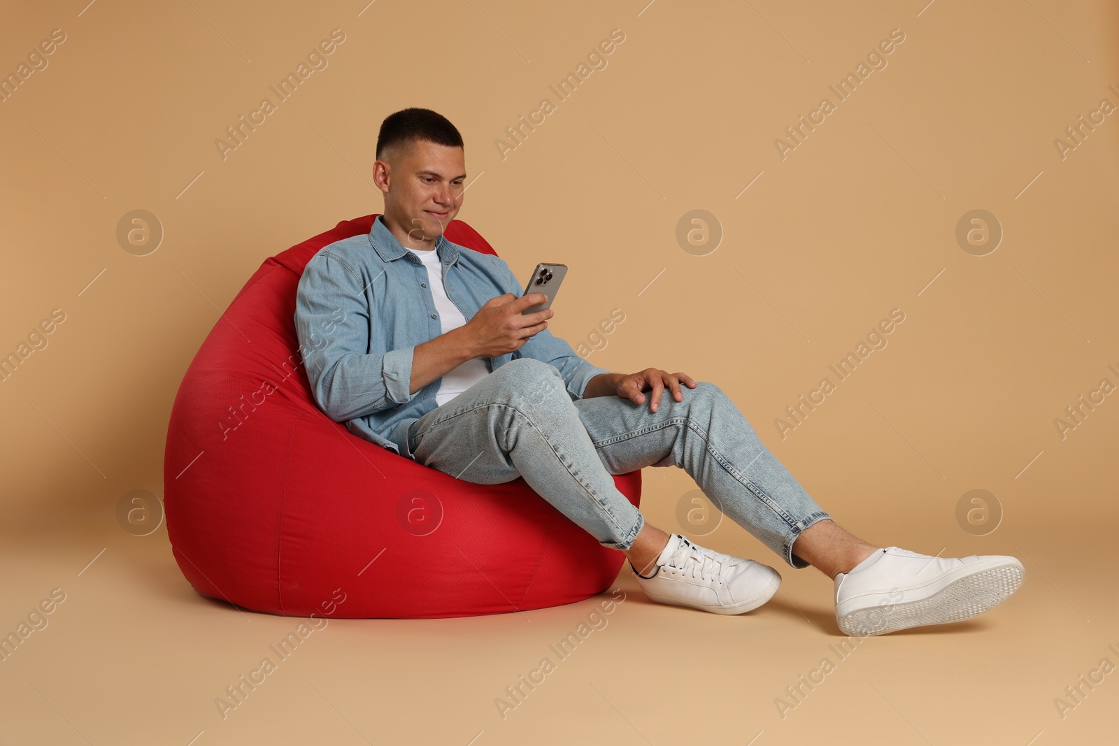 Photo of Handsome man with smartphone on red bean bag chair against beige background