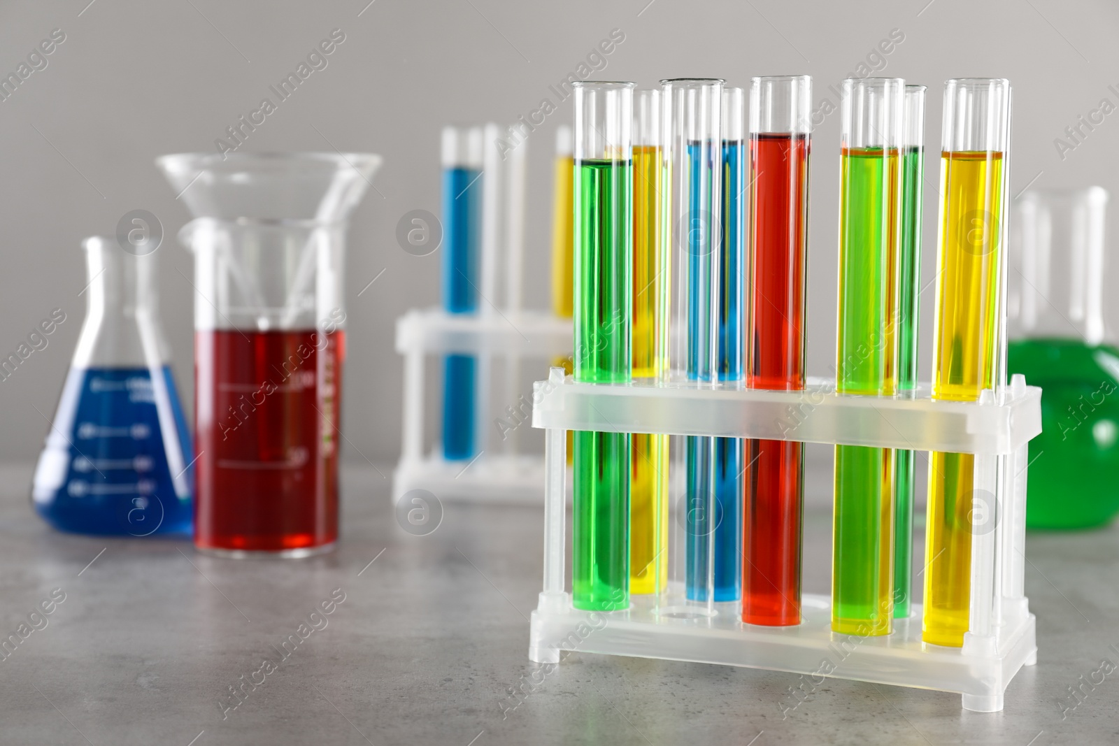Photo of Test tubes with liquids in stand and flasks on table against light grey background, selective focus