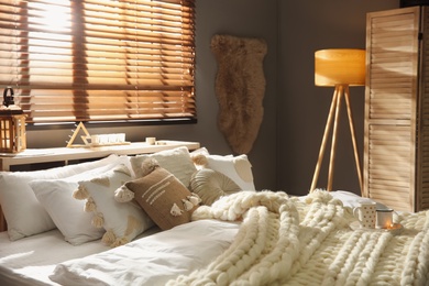 Bed with cozy knitted blanket and cushions near window in room. Interior design