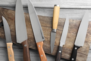 Photo of Many different knives and board on light wooden table, flat lay