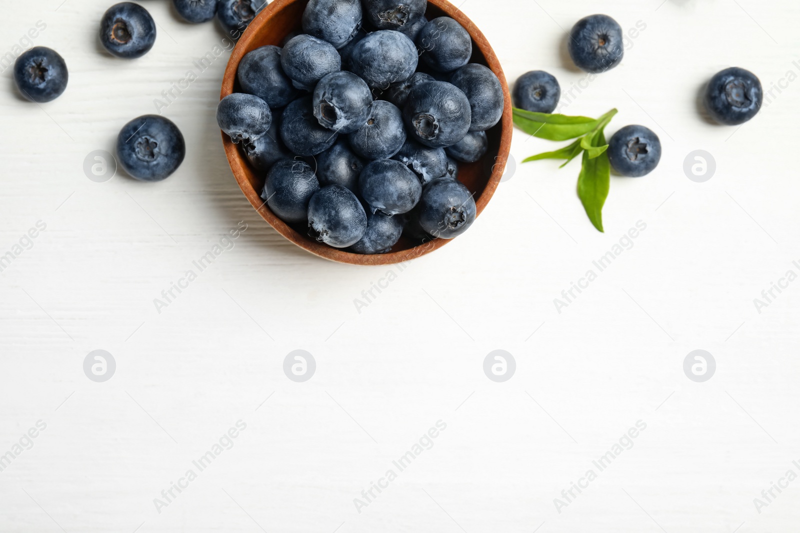 Photo of Fresh ripe blueberries in bowl on white wooden table, flat lay. Space for text